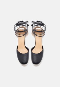 Paire d'espadrilles plates à bout fermé noires avec semelles intérieures caramel et lanières noires autour de la cheville sur fond blanc.