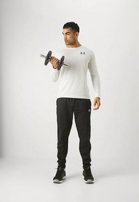 Fehér hosszú ujjú ingben és fekete nadrágban álló férfi, aki jobb karjával súlyzót emel, egyszerű, világos háttér előtt.