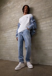 T-shirt bianca con logo, giacca di jeans azzurro chiaro e jeans azzurro chiaro. Sneaker beige con dettagli. Sfondo in cemento spazzolato.
