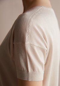 Lys beige kortærmet sweater med en glat tekstur og ribbede manchetter. Har en subtil søm langs skulderen for et tætsiddende udseende.