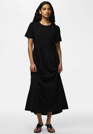 Femme aux cheveux bouclés foncés portant une robe midi noire à manches courtes et des sandales à talons ouvertes noires, debout devant un fond uni.