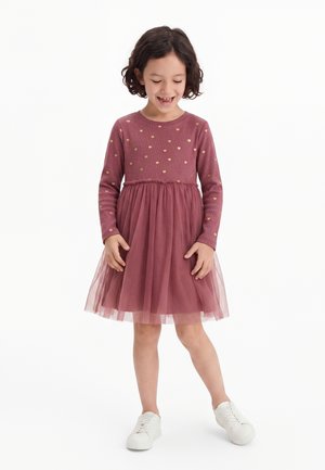 Jeune fille souriante, vêtue d'une robe rose à manches longues avec des motifs de cœurs dorés et des baskets blanches, debout sur un fond blanc.