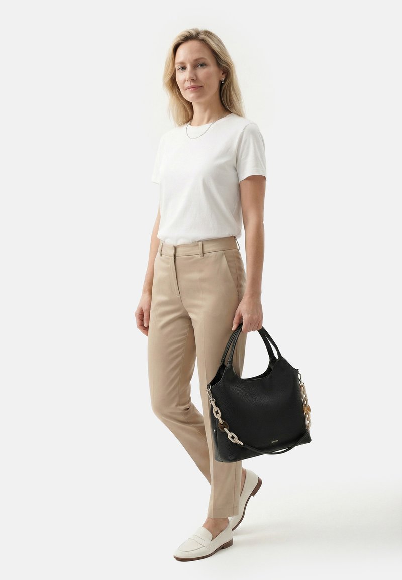 Woman wearing white t-shirt, beige pants, and white loafers holding black handbag with chain strap, standing against plain white background.