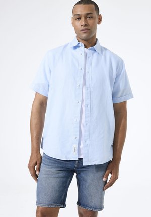 Homme portant une chemise bleu clair à manches courtes boutonnée par-dessus un t-shirt blanc et un short en jean bleu, debout devant un fond blanc.