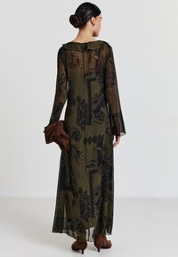 Robe longue en voile d'olive avec un motif paisley noir, décolleté froncé et longues manches évasées. Le mannequin tient une pochette en tissu marron.