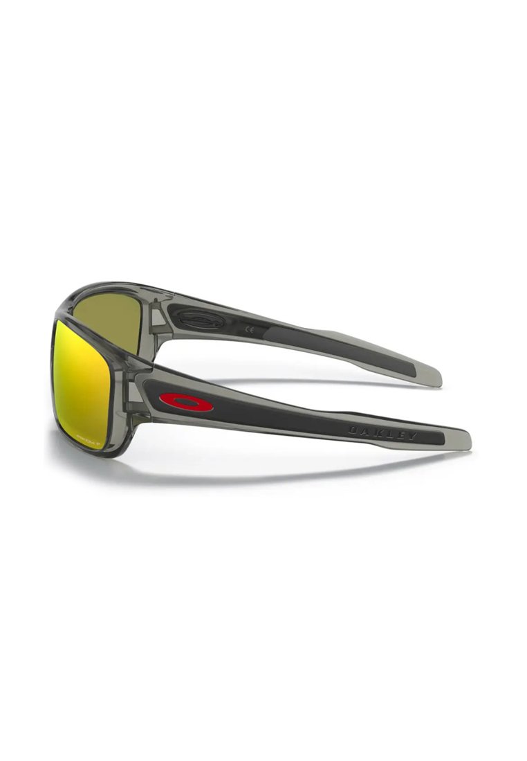 Gafas de sol Oakley grises con lentes reflectantes amarillas y logo rojo en la patilla, mostradas desde un ángulo lateral sobre un fondo blanco.