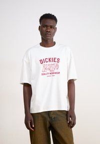 T-shirt bianco in cotone con maniche corte e collo rotondo, caratterizzato da una stampa grafica rossa che include il testo "Dickies Quality Workwear".