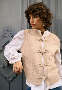 Béžová pletená vesta s vrkočovým vzorom a guľatými detailmi, so zapínaním na gombíky vpredu. Nosená cez bielu, volánikovú košeľu s dlhými rukávmi.