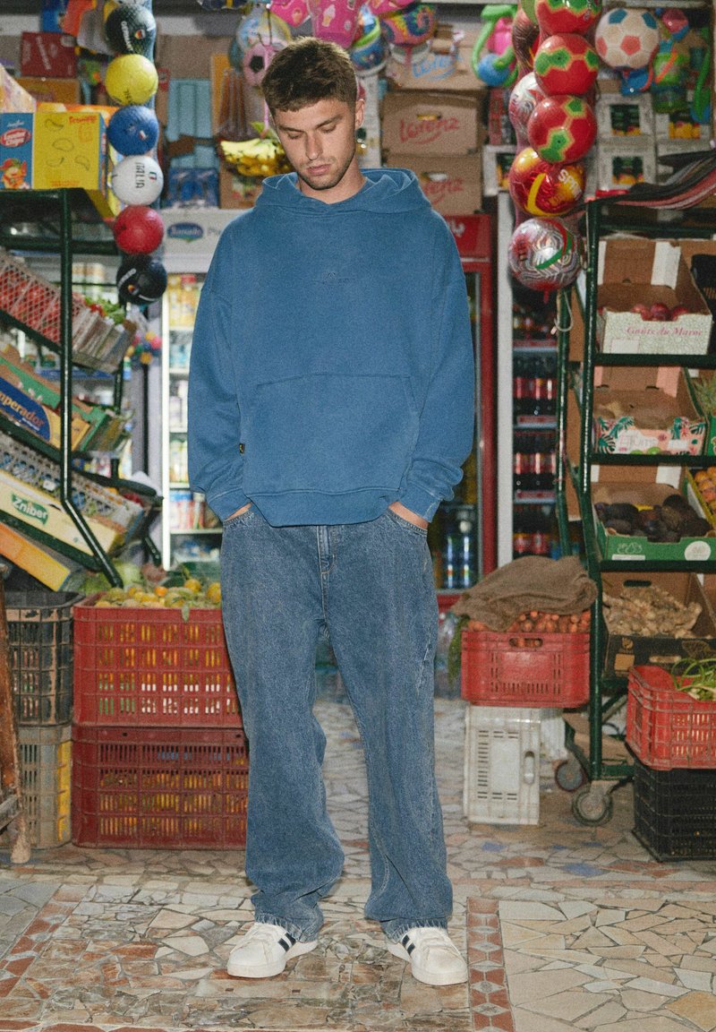 Młody mężczyzna w niebieskiej bluzie z kapturem i dżinsach stoi w zatłoczonym targu wśród poustawianych skrzyń, wiszących piłek i półek z towarami.