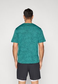 Αθλητική μπλούζα σε τιρκουάζ απόχρωση με υφή, κοντά μανίκια, στρογγυλή λαιμόκοψη και διακριτική λεπτομέρεια λογότυπου στο πίσω μέρος. Συνδυάζεται με μαύρο σορτς.