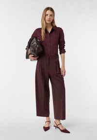 Burgundowa koszula z długim rękawem zapinana na guziki, dopasowane szerokie spodnie, w zestawieniu z burgundowymi butami na obcasie typu slingback oraz brązową torbą.