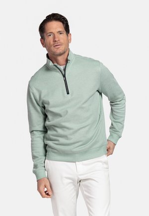 Mann trägt einen hellgrünen Pullover mit Reißverschluss bis zur Brust und weiße Hose, steht vor einem schlichten weißen Hintergrund, mit einer Hand in der Tasche.