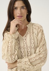 Beige lace blouse with a V-neck, puffed sleeves, and intricate circular patterns. Delicate texture with ruffled collar and cuffs.