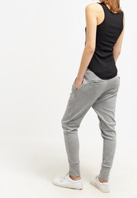 Kobieta stojąca z rękami w kieszeniach, ubrana w czarny top na ramiączkach, szare spodnie dresowe i białe sneakersy, odwrócona plecami na białym tle.