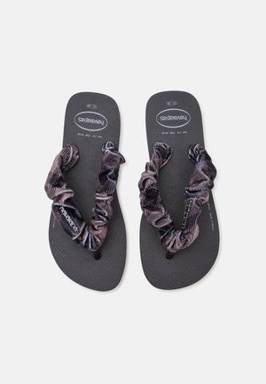 Ζευγάρι μαύρα σαγιονάρες Havaianas με σκούρες ύφαντες λουρίδες με πιέτες, τοποθετημένες επίπεδα με ορατό κείμενο στη σόλα σε ανοιχτό γκρι φόντο.