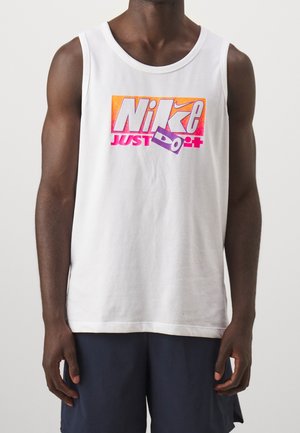 Person, die ein weißes ärmelloses Shirt mit einem bunten "Nike Just Do It"-Logo und dunkle Shorts vor einem schlichten Hintergrund trägt.