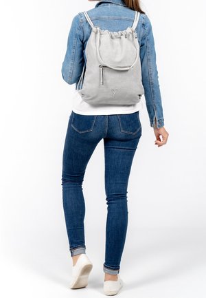 Un sac à dos en maille gris avec un cordon de serrage et une poche zippée à l'avant, reposant sur le dos d'une personne portant une veste en denim et un jeans.