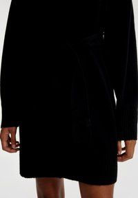 Schwarzes, gestricktes Kleid mit langen Ärmeln, einem Bindegürtel in der Taille und gerippten Ärmelabschlüssen; knielang mit einer weichen Textur.