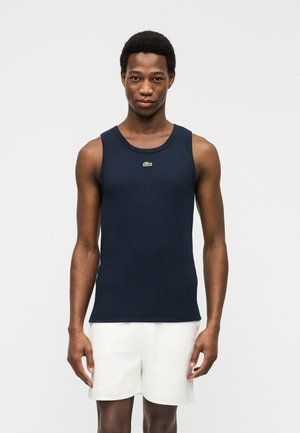 Młody mężczyzna stojący twarzą do przodu, ubrany w granatową koszulkę bez rękawów z małym logo krokodyla oraz białe spodenki.