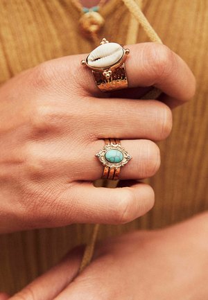 Bagues en or avec des textures distinctes ; l'une présente une coquille, l'autre une pierre turquoise. Une main les tenant montre la texture de la peau et des tons chauds.