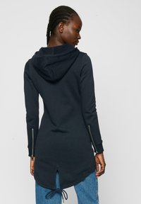 Hoodie long bleu marine en tissu doux. Il présente un ourlet à cordon de serrage, des poches zippées sur les côtés et un détail fendu dans le dos pour la ventilation.