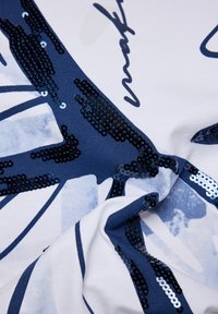 Baumwollstoff mit weißem Grund, der ein Design in Marineblau und Pailletten als Akzent aufweist. Texturiert, mit einer glatten Oberfläche und nahtloser Integration der Elemente.