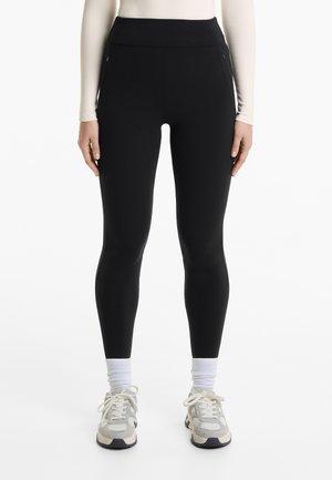 Zwarte high-waisted leggings van rekbaar materiaal met zijzakken, gecombineerd met grijs-witte sneakers en witte enkelsokken.