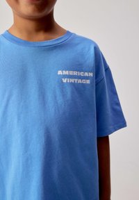 Enfant portant un T-shirt bleu clair avec le texte blanc "AMERICAN VINTAGE" sur la poitrine gauche, sur un fond clair uni.