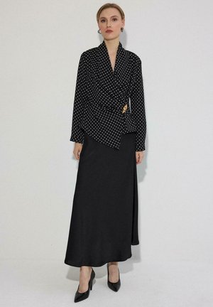 Nő fekete pöttyös átkötős blúzban arany csattal és hosszú fekete szoknyában, fekete hegyes orrú magas sarkú cipővel.