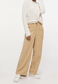 Pantalon beige à jambes larges avec taille à cordon et poches avant, associé à un haut blanc texturé à manches longues et des baskets blanches.
