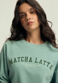 Miętowy sweter z okrągłym dekoltem, z napisem "MATCHA LATTE" w ciemnozielonym kolorze oraz mniejszym hasłem w białym kolorze. Miękka tekstura materiału.