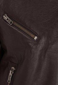 Giacca in pelle marrone con zip. Presenta una piccola tasca sul petto e una tasca laterale più grande, evidenziando una texture liscia e un colore ricco.