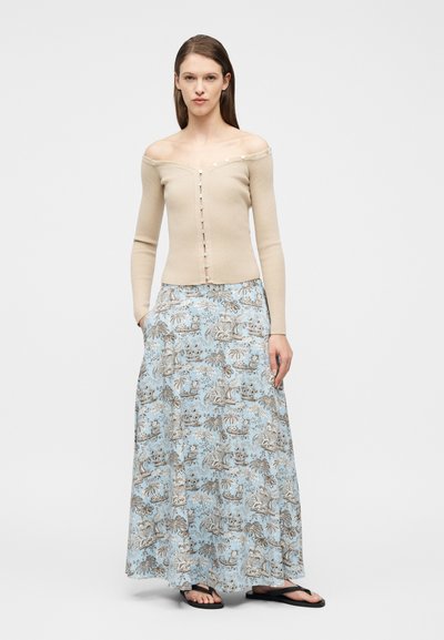 Γυναίκα που φοράει μπεζ πουλόβερ με κουμπιά και ανοιχτούς ώμους, μπλε μακριά φλοράλ φούστα με τσέπες και μαύρα σαγιονάρες, στέκεται μπροστά σε ένα απλό φόντο.