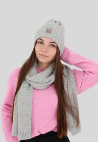 Cappello a coste grigio chiaro con un patch del brand, abbinato a una sciarpa coordinata, indossato sopra un maglione rosa, su uno sfondo neutro.