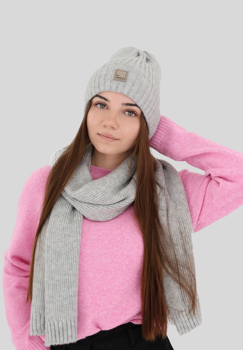 Cappello a coste grigio chiaro con un patch del brand, abbinato a una sciarpa coordinata, indossato sopra un maglione rosa, su uno sfondo neutro.