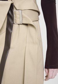 Jupe beige avec une boucle de ceinture rectangulaire argentée et une bordure verticale marron foncé, portée avec un haut marron foncé à manches longues et une main visible.