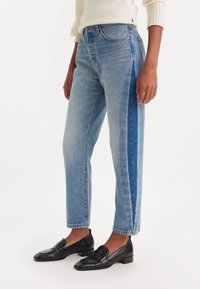 Vaqueros de denim azul claro con un corte recto, que cuentan con paneles laterales en azul más oscuro. Combinados con mocasines de cuero negros.