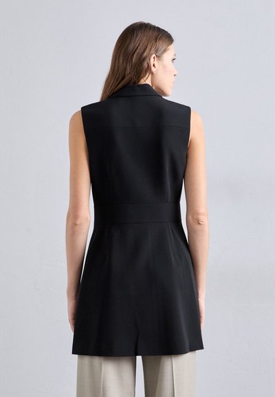 Melnā bezpiedurkņu blūze ar pieguļošu jostasvietu un garāku aizmugurējo apdari. Šūšanas dizains izceļas ar tīrām līnijām un minimālistisku struktūru.