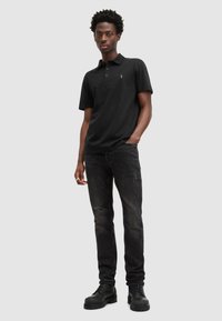 Schwarzes Polohemd mit Kragen und drei Knöpfen, kombiniert mit dunklen Jeans. Das Outfit wird mit schwarzen Kampfstiefeln abgerundet.