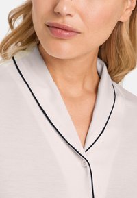 Ljusgrå blus med sjalkrage, marinblå piping och knappstängning framtill, tillverkad av mjukt tyg som framhäver en slät yta.