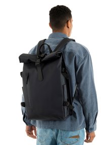 Μαύρο αδιάβροχο σακίδιο πλάτης με ανθεκτικό πάνω κύλινδρο, ρυθμιζόμενους ιμάντες, μπροστινή τσέπη με φερμουάρ και κομψό σχεδιασμό, εμφανιζόμενο σε άτομο.