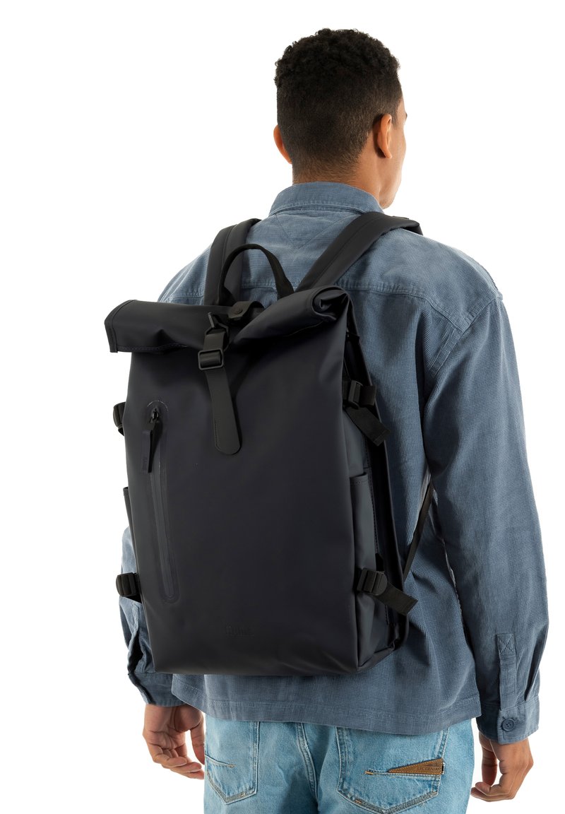 Μαύρο αδιάβροχο σακίδιο πλάτης με αν θεκτικό πάνω κύλινδρο, ρυθμιζόμενους ιμάντες, μπροστινή τσέπη με φερμουάρ και κομψό σχεδιασμό, εμφανιζόμενο σε άτομο.