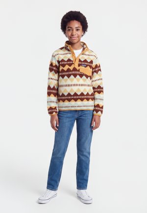 Ung person iført en mønstret pullover med geometriske former, blå jeans og hvide sneakers på en hvid baggrund.
