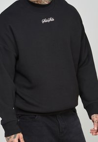 Felpa nera in misto cotone, con collo rotondo e polsini a costine. Sul davanti è presente un testo ricamato bianco con il logo "SikSilk".
