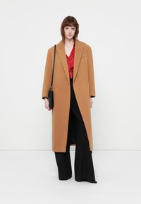 Palton oversized de culoare cămilă, cu un design structurat, având o singură buzunar și rever late, purtat peste un top roșu și pantaloni negri.