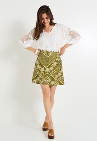 Blusa blanca con detalles de encaje y cordones, falda envolvente verde con estampado de palmeras, combinada con sandalias de plataforma marrones.