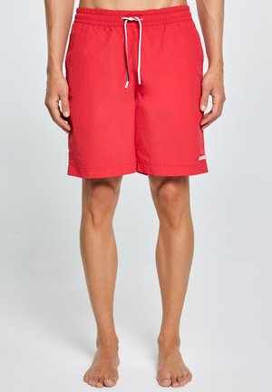Czerwone męskie spodenki kąpielowe ze białym sznurkiem, elastycznym pasem i logo przy krawędzi, noszone boso na jednolitym tle.