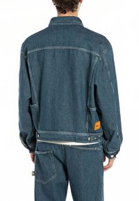 Denim jacket in dark blue, featuring a collared neck, long sleeves, and an orange label on the back. Includes side pockets and a zipper.