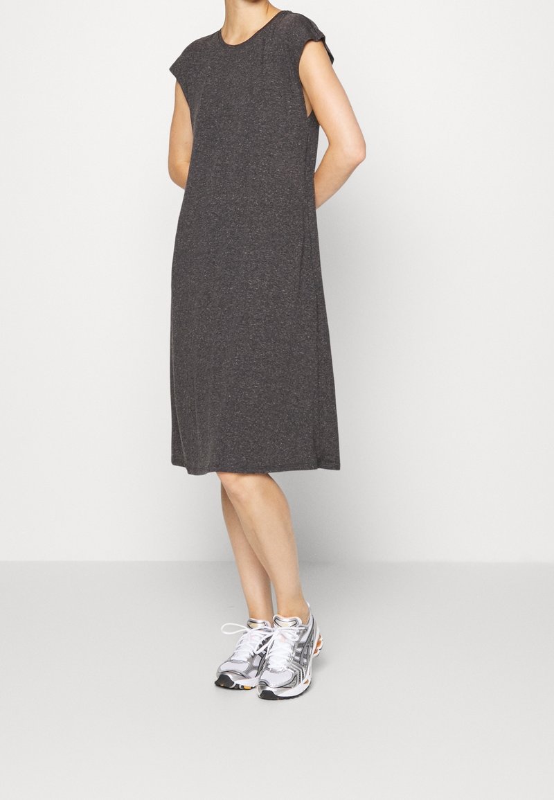 Personne portant une robe ample, gris foncé, sans manches et arrivant au genou, avec des baskets argentées, debout devant un fond clair et uni.