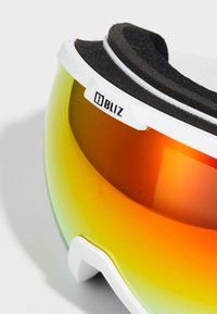 Ski brilles ar baltu rāmīti, oranži-dzeltenu spoguļa lēcu un melnu putu polsterējumu iekšpusē komfortam. Logo sānos.